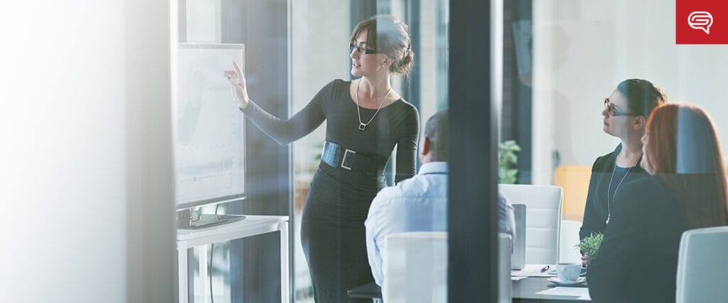 A woman in professional attire stands in a bright conference room, highlighting a slide focused on client experience for consulting firms on a large screen. Three colleagues, seated at a table, are engaged with the presentation. The room is modern with large glass windows.